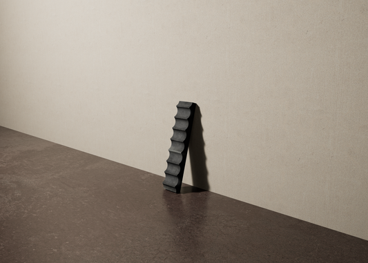 a 2"x10" bespoke fluted concrete tile with eight peaks leans against a textured, plaster wall on an unfinished concrete floor